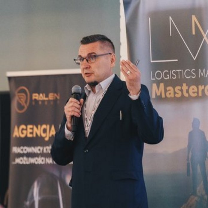 mężczyzna przemawiający przed publicznością, a za nim standy